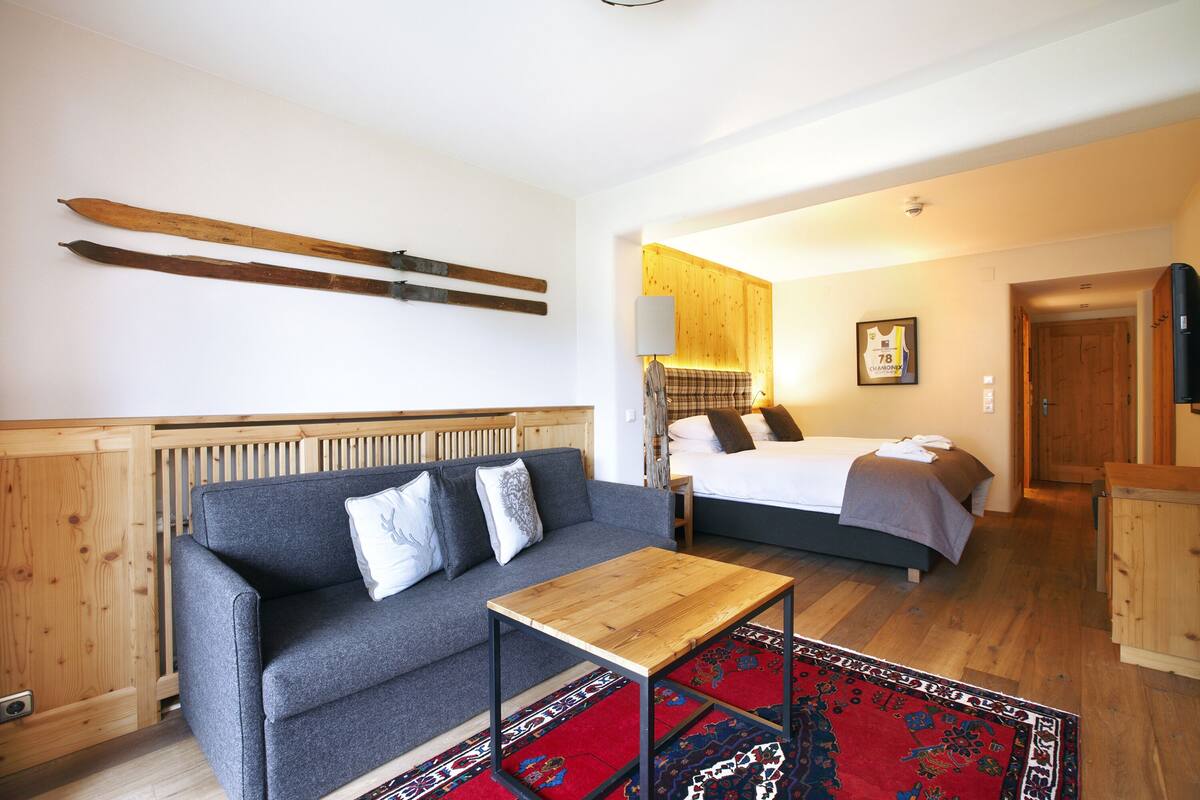 Una habitación con un sofá gris, una mesa de madera, una cama con un tapete rojo y azul estampado, y paredes de madera.