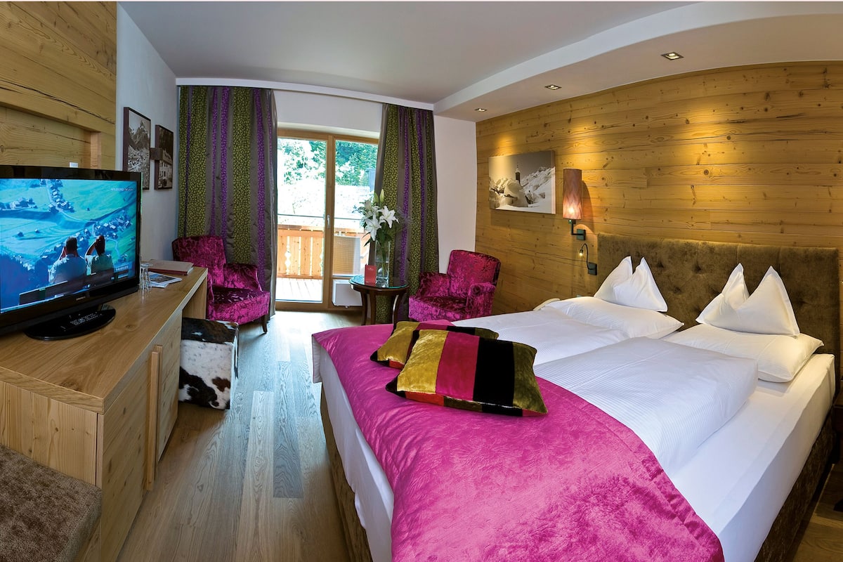 Habitación de hotel con pared de madera, televisor, cama con ropa de cama rosa, una silla y una mesita.