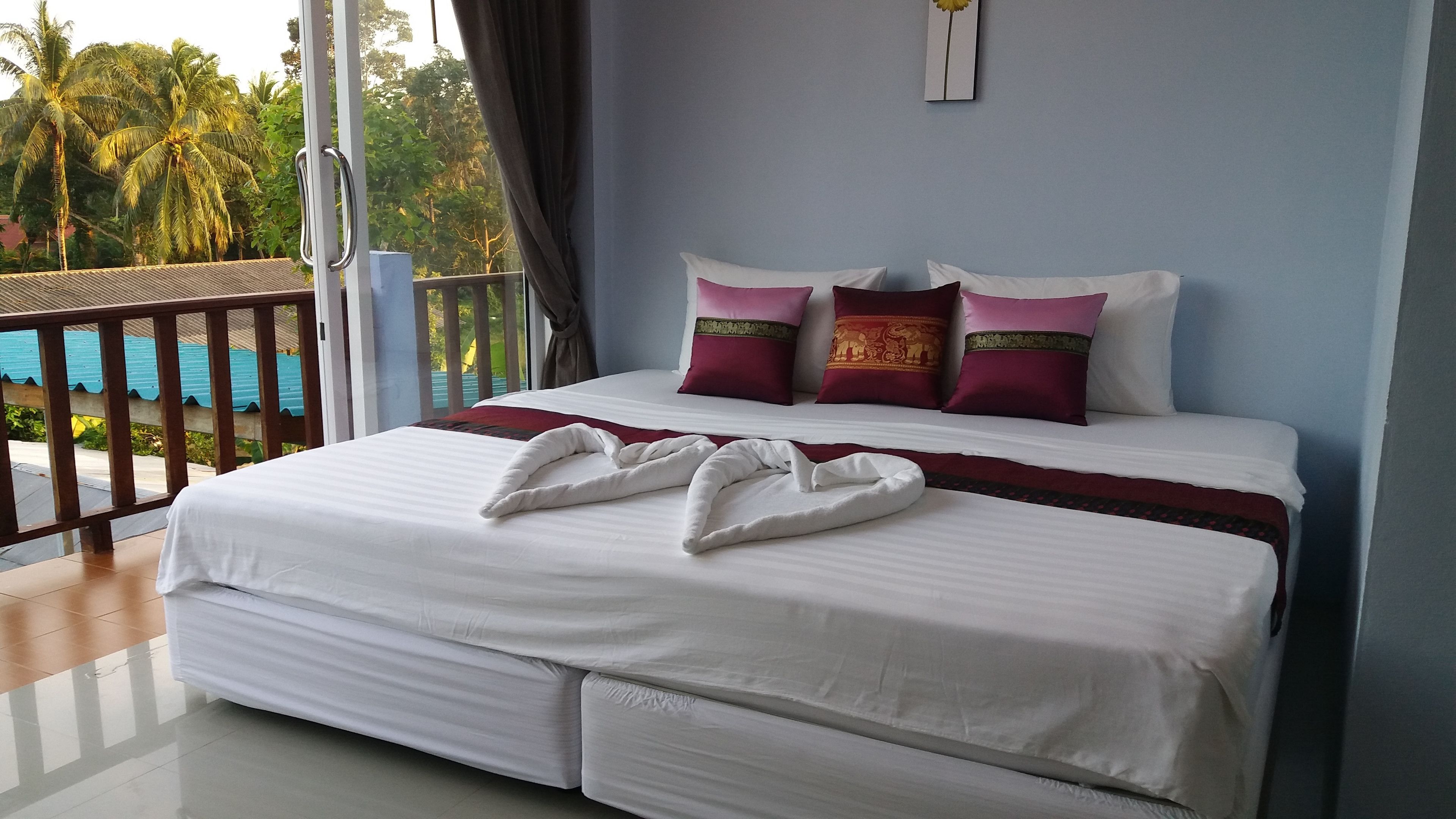 A neatly made bed with white linens and decorative pillows, a balcony with a view of palm trees and a pool, and a blue wall.