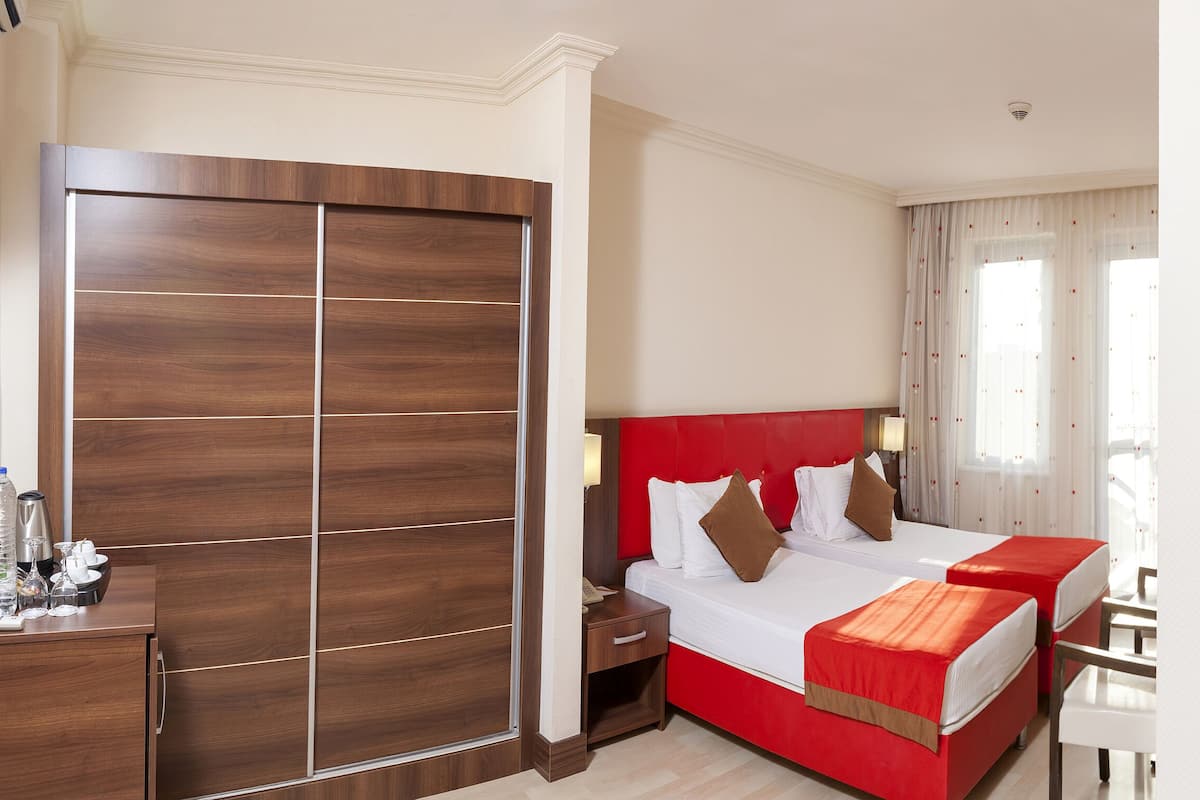 A hotel room with a large wooden wardrobe, a red bed with white bedding, a bedside table with a lamp, and a desk with a chair.