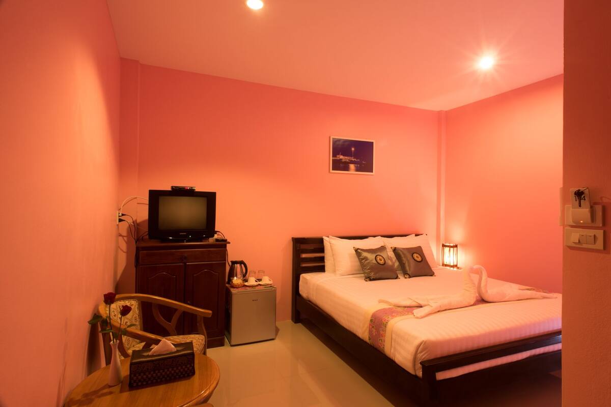 A bedroom with a bed, a television on a dresser, a small table with a vase and a tissue box, and a nightstand with a lamp.