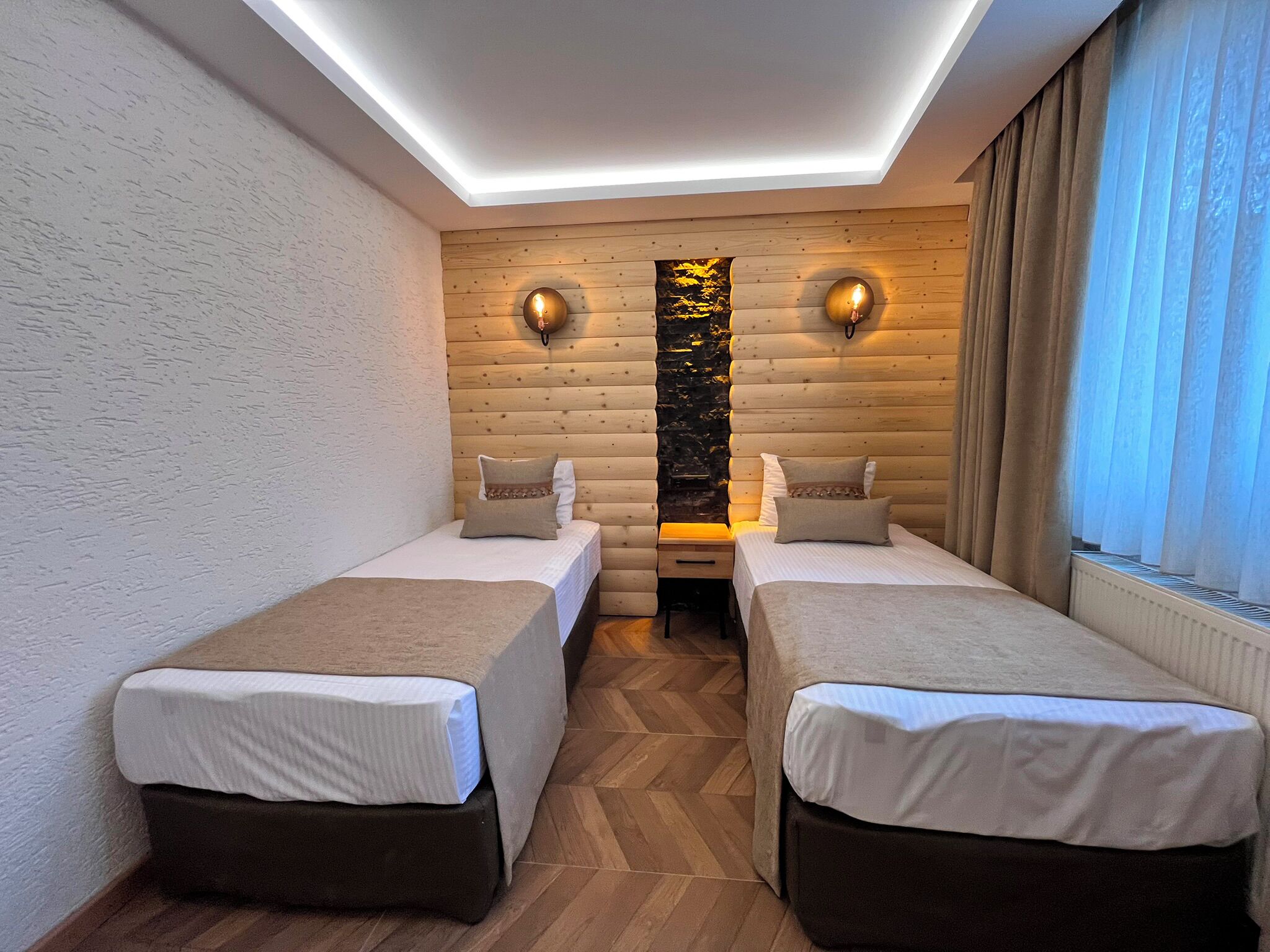 A modern room with two beds, wooden panel walls, a stone accent wall, and a window with curtains.