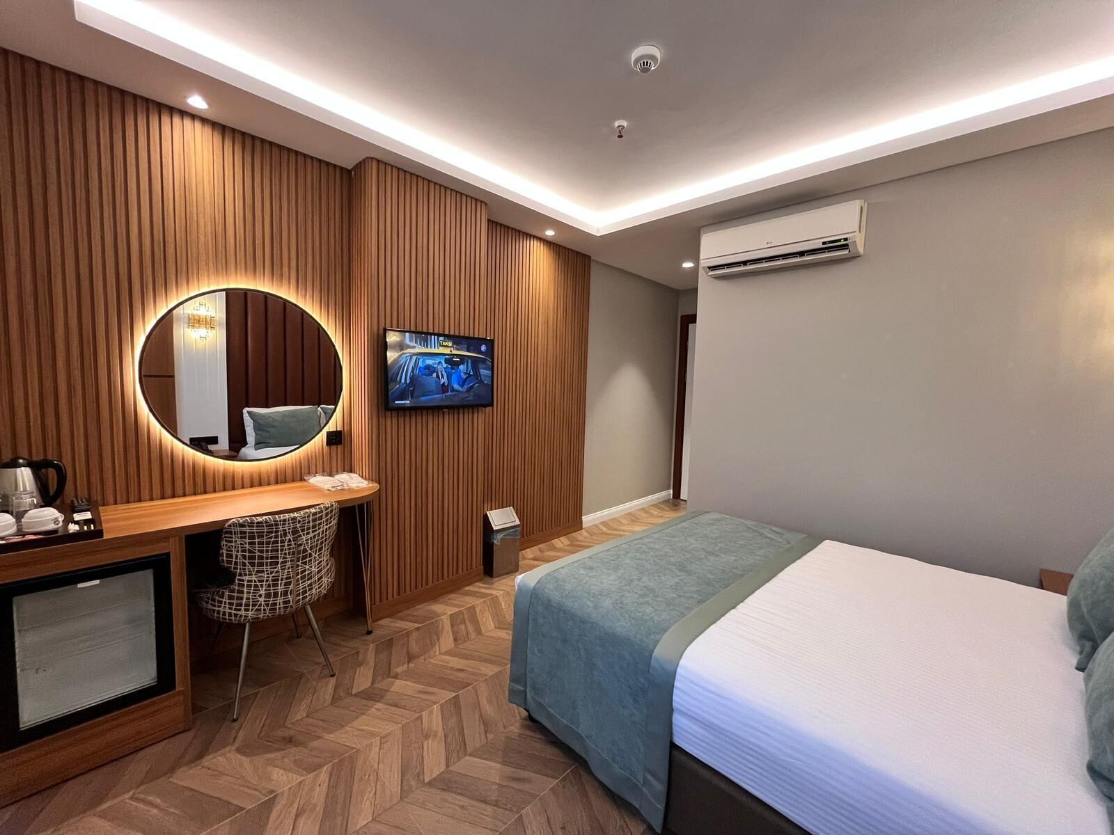 A modern hotel room with a wooden panel wall, a round mirror, a flat-screen TV, a bed with a blue bedspread, and a desk with a kettle and a basket.