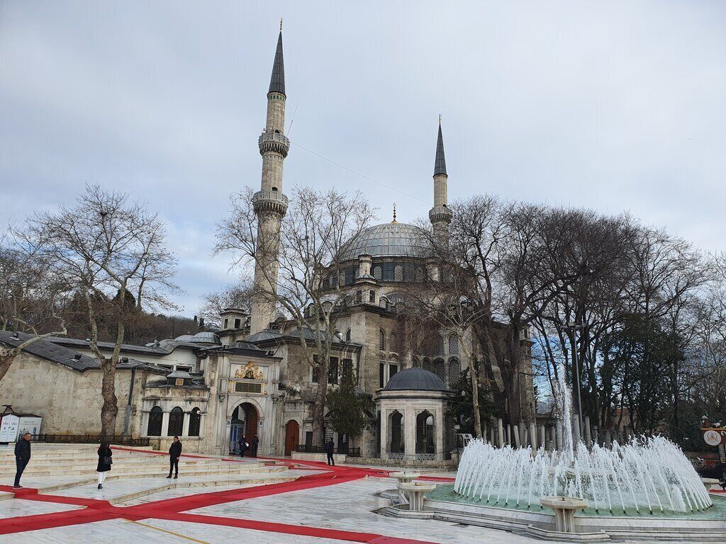 A historic building with two minarets and a central dome, surrounded by a fountain and a red carpeted area.