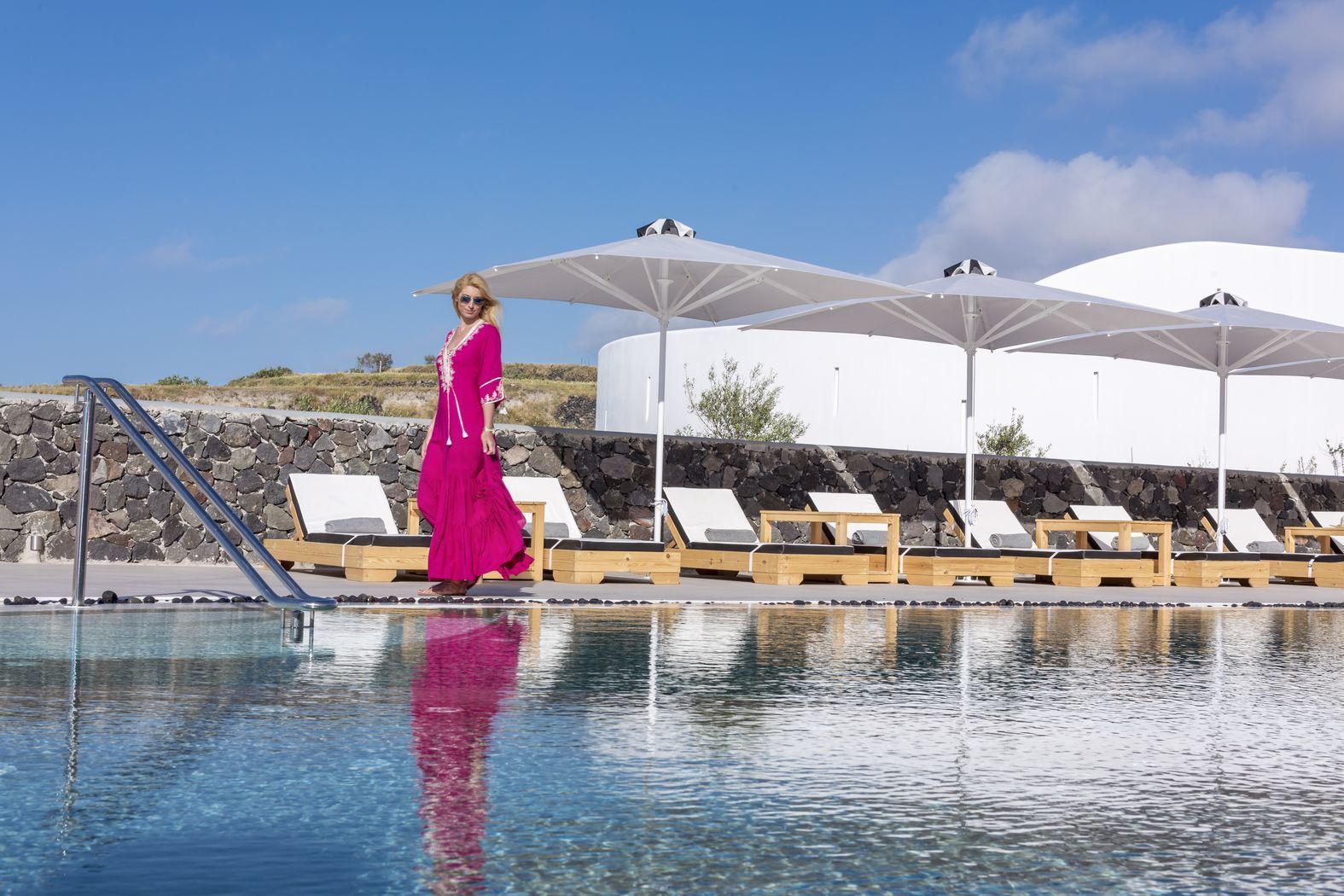 Un espace piscine avec des chaises longues et des parasols, un mur en pierre et un ciel dégagé.