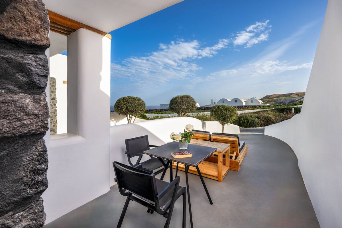 A modern outdoor seating area with a table and chairs, a stone wall, and a view of white buildings and trees under a clear sky.