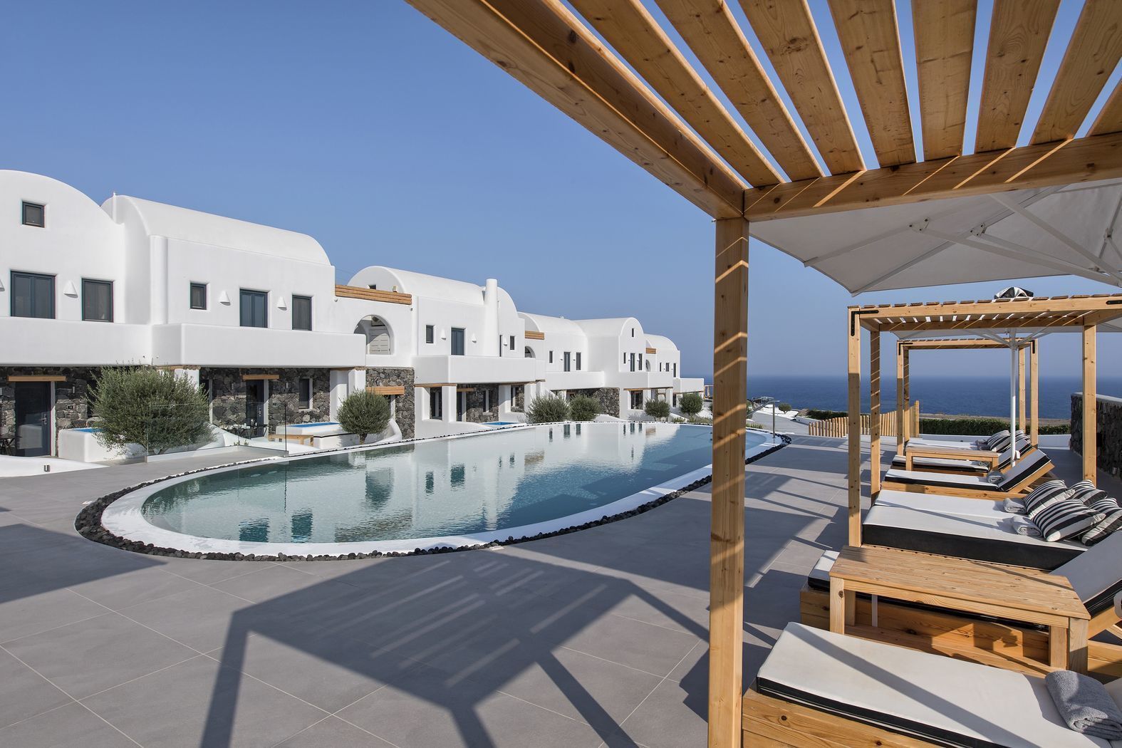 A modern resort with a large infinity pool, white buildings, and a wooden pergola with lounge chairs.