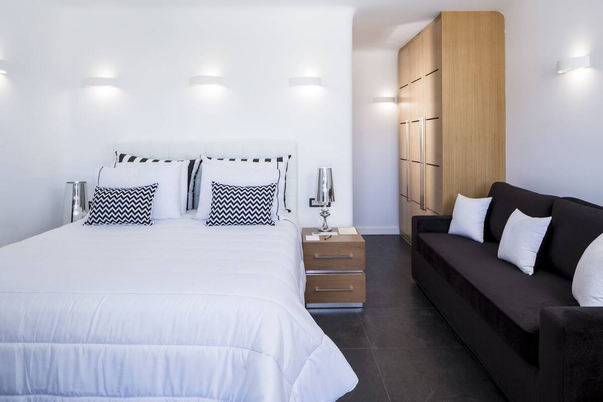A modern bedroom with a large bed, bedside tables, a dark sofa, and built-in wooden wardrobe.