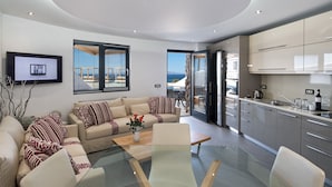 Una sala de estar moderna con una mesa de centro de vidrio, sofá color beige con cojines estampados, televisor de pantalla plana y vistas al exterior a través de grandes ventanales.