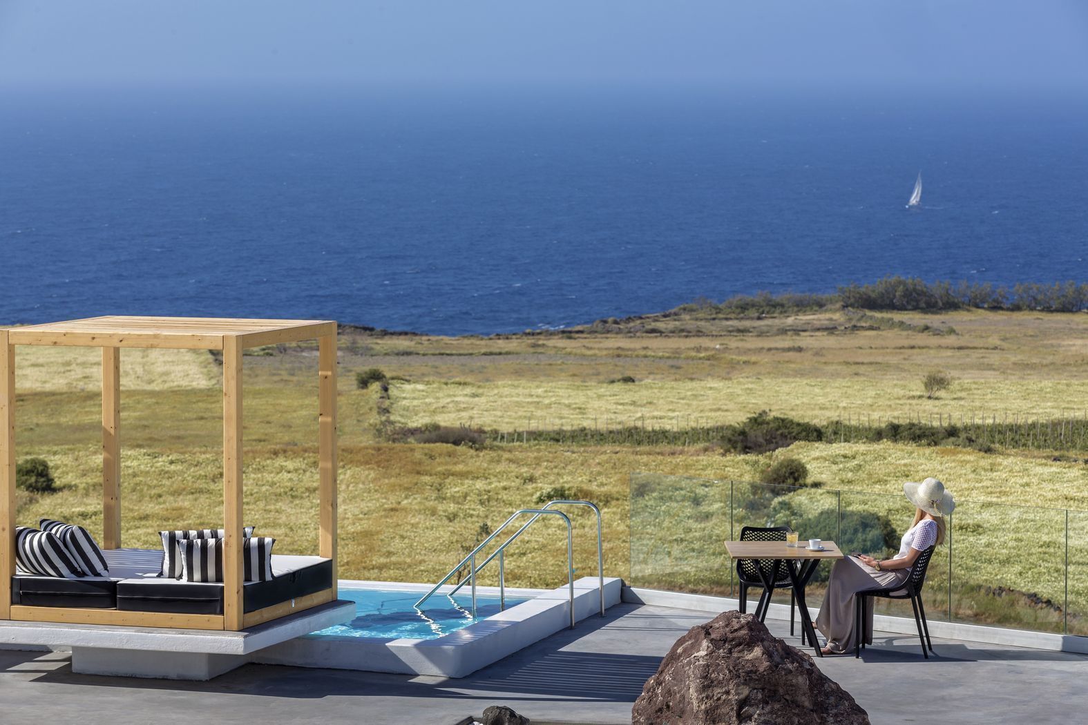 Un área de piscina con cubierta de madera, sillas de descanso y una mesa donde se sienta una persona.