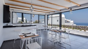Un comedor moderno con sillas y mesas blancas, grandes ventanales que ofrecen vista al mar y edificios blancos.