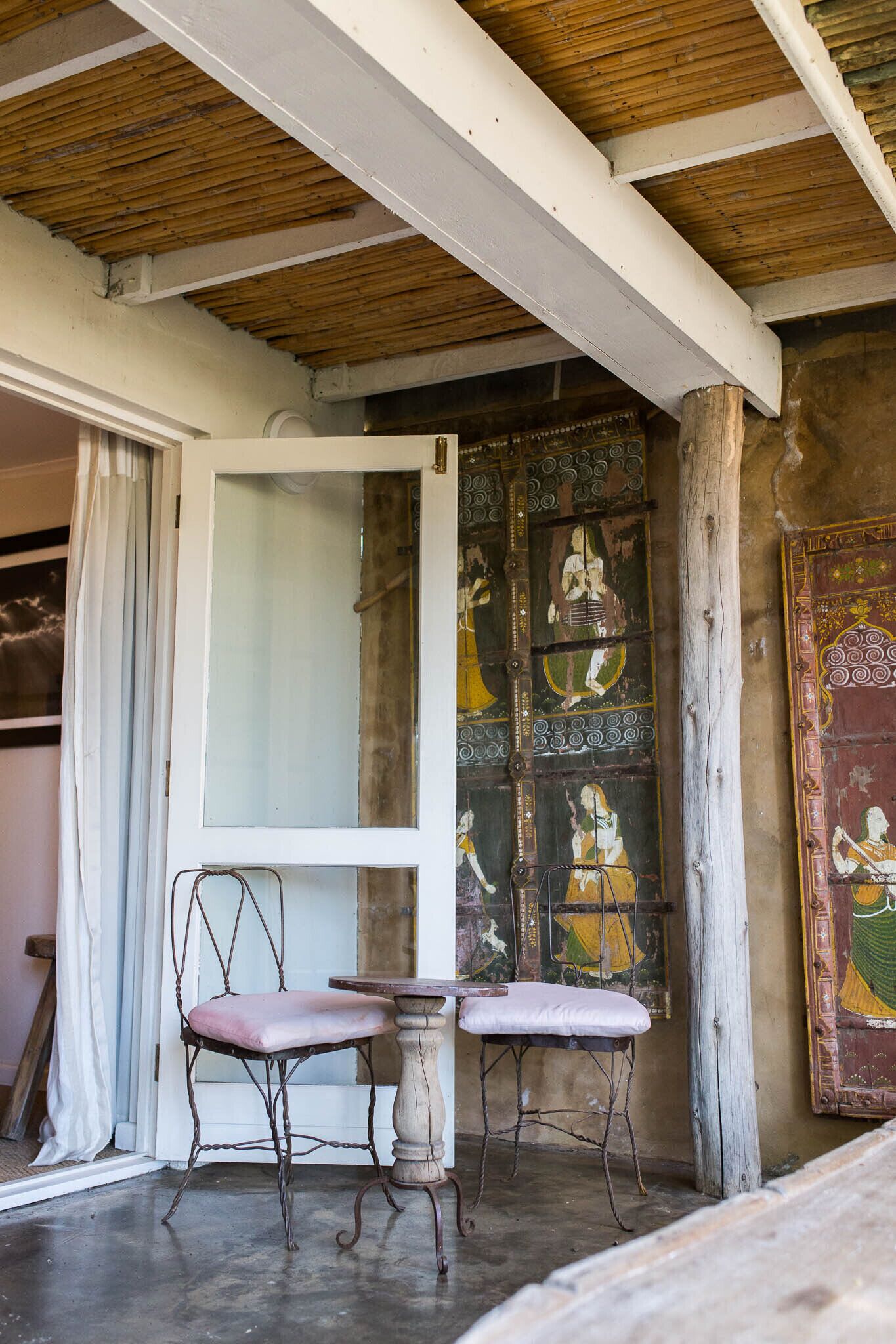 A room with a wooden ceiling, a white door, and a wall with colorful artwork. There are two chairs and a small table.