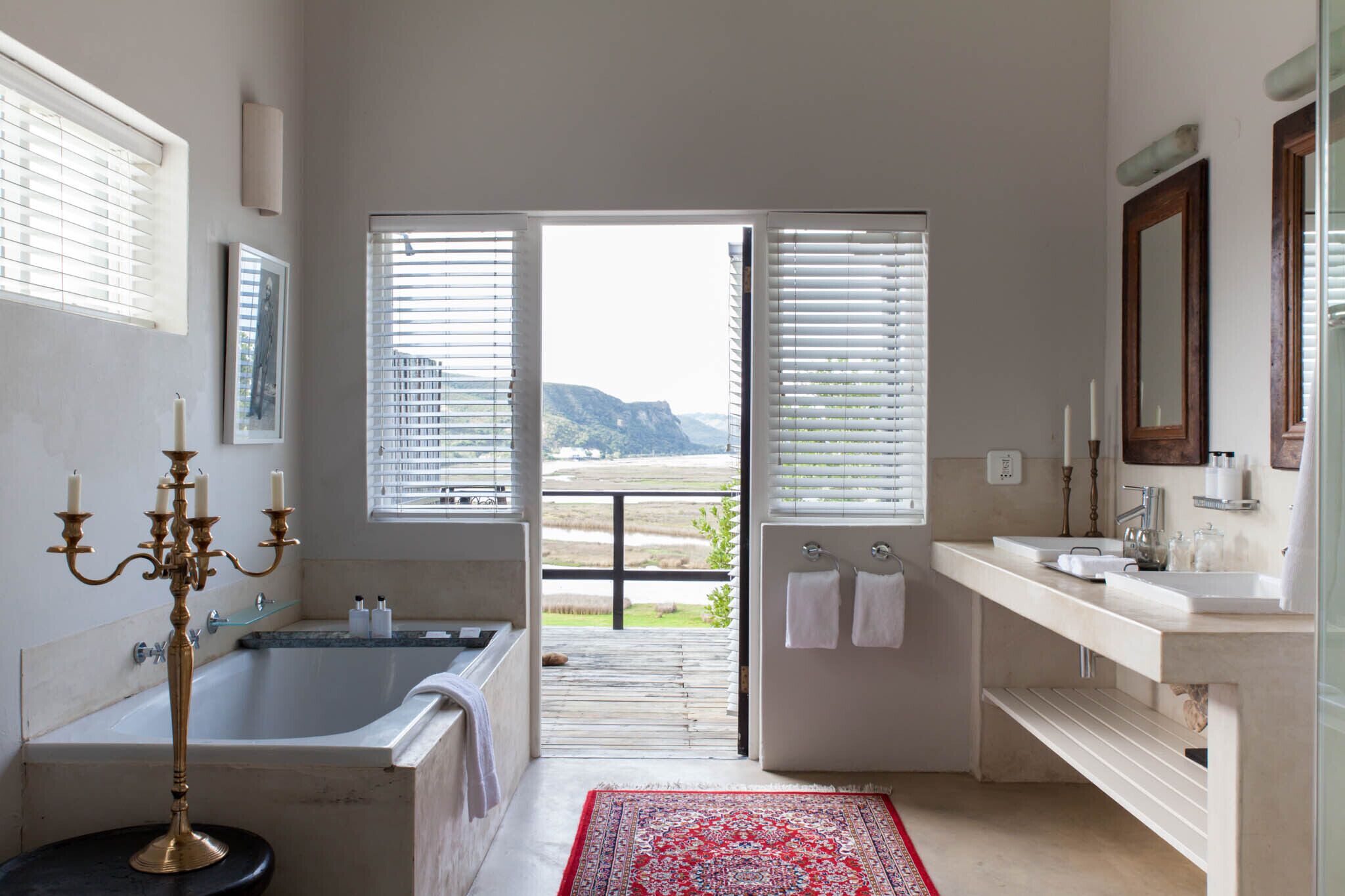 A bathroom with a bathtub, a large mirror, and a view of a mountainous landscape through open windows.