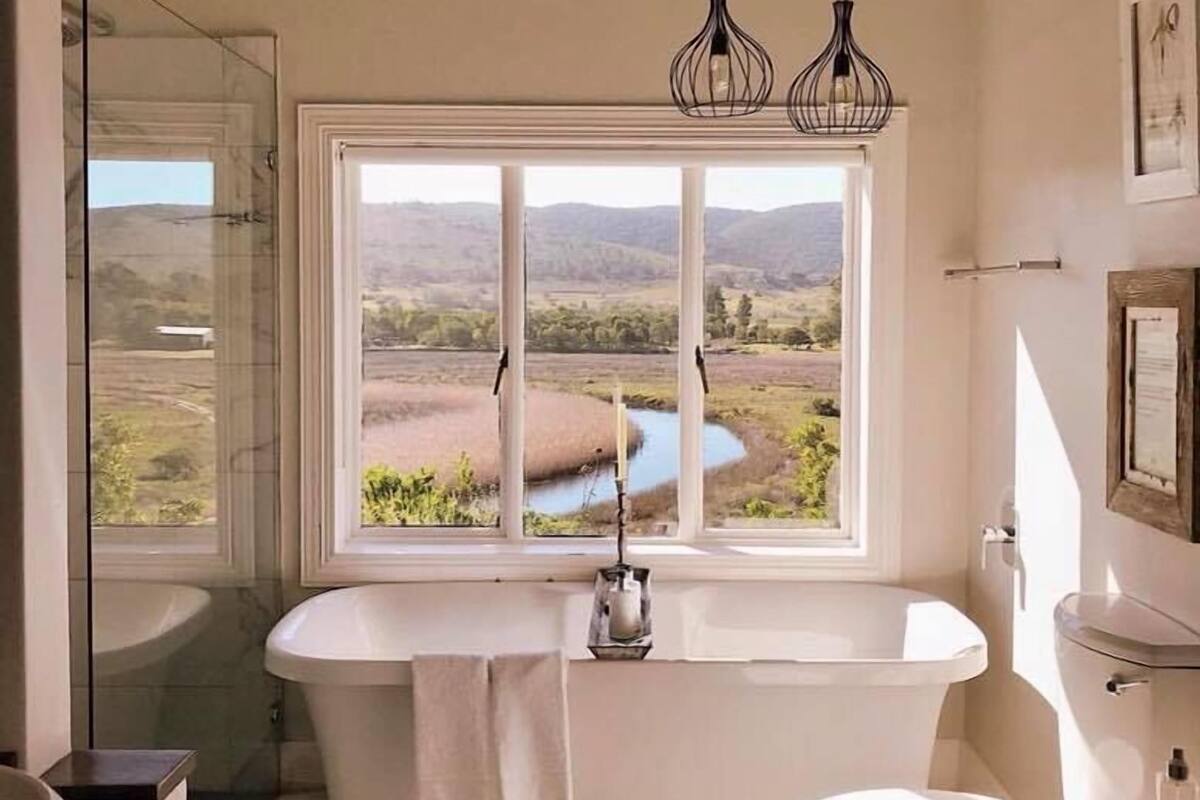 Une salle de bain équipée d’une baignoire, de toilettes, d’un lavabo et d’une grande fenêtre donnant sur un paysage.