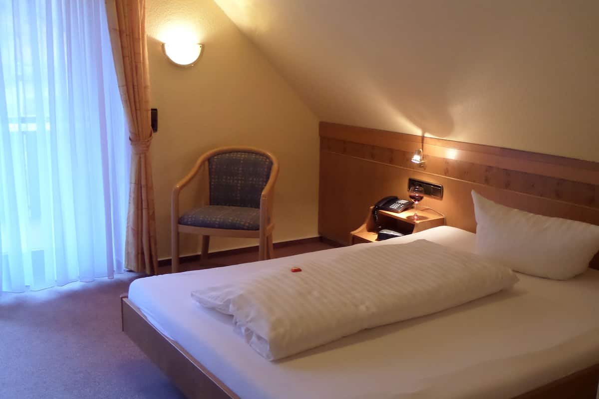 A hotel room with a bed, a chair, a bedside table with a phone and a glass of wine, and a wall-mounted light.
