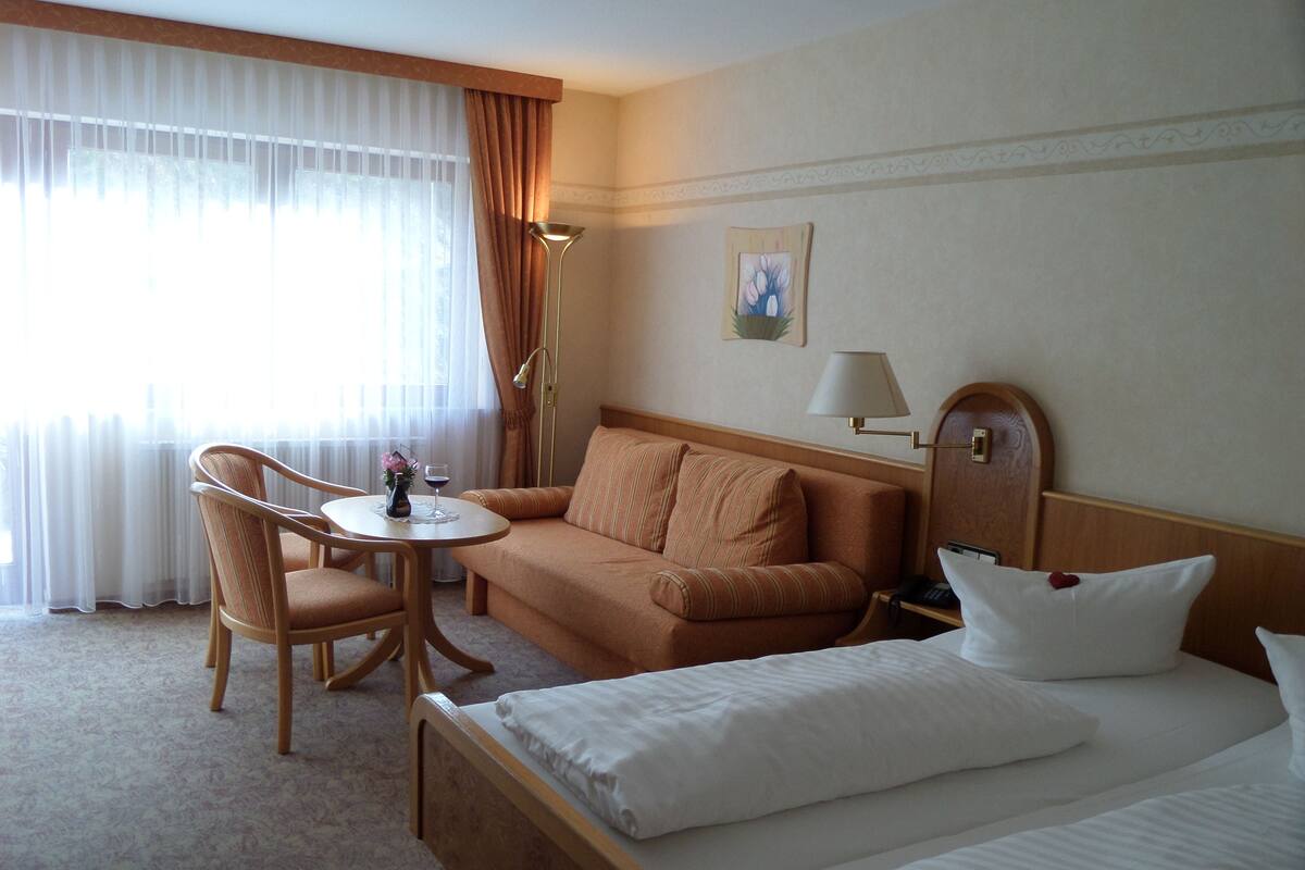 A hotel room with a bed, a sofa, a chair, a small table with a vase and a glass of wine, and a lamp.