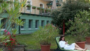 Una persona descansando en una silla verde en un patio con plantas en macetas y un edificio con balcones al fondo.