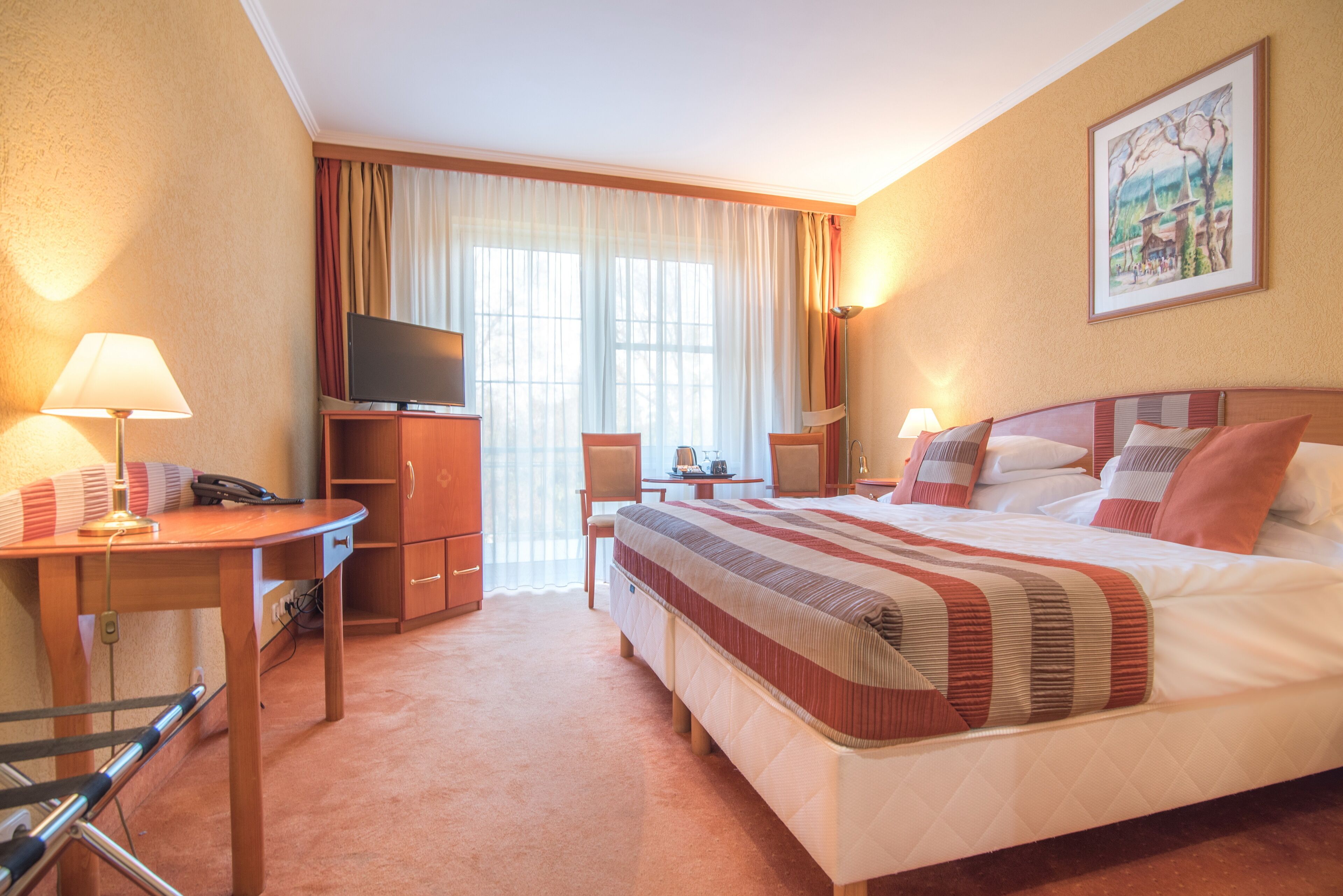 Habitación de hotel con cama, escritorio con lámpara, televisión, silla y ventana con cortinas.