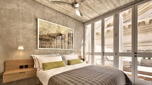 Une chambre avec un grand lit, une table de chevet en bois, un ventilateur de plafond et une grande fenêtre donnant sur le paysage urbain.