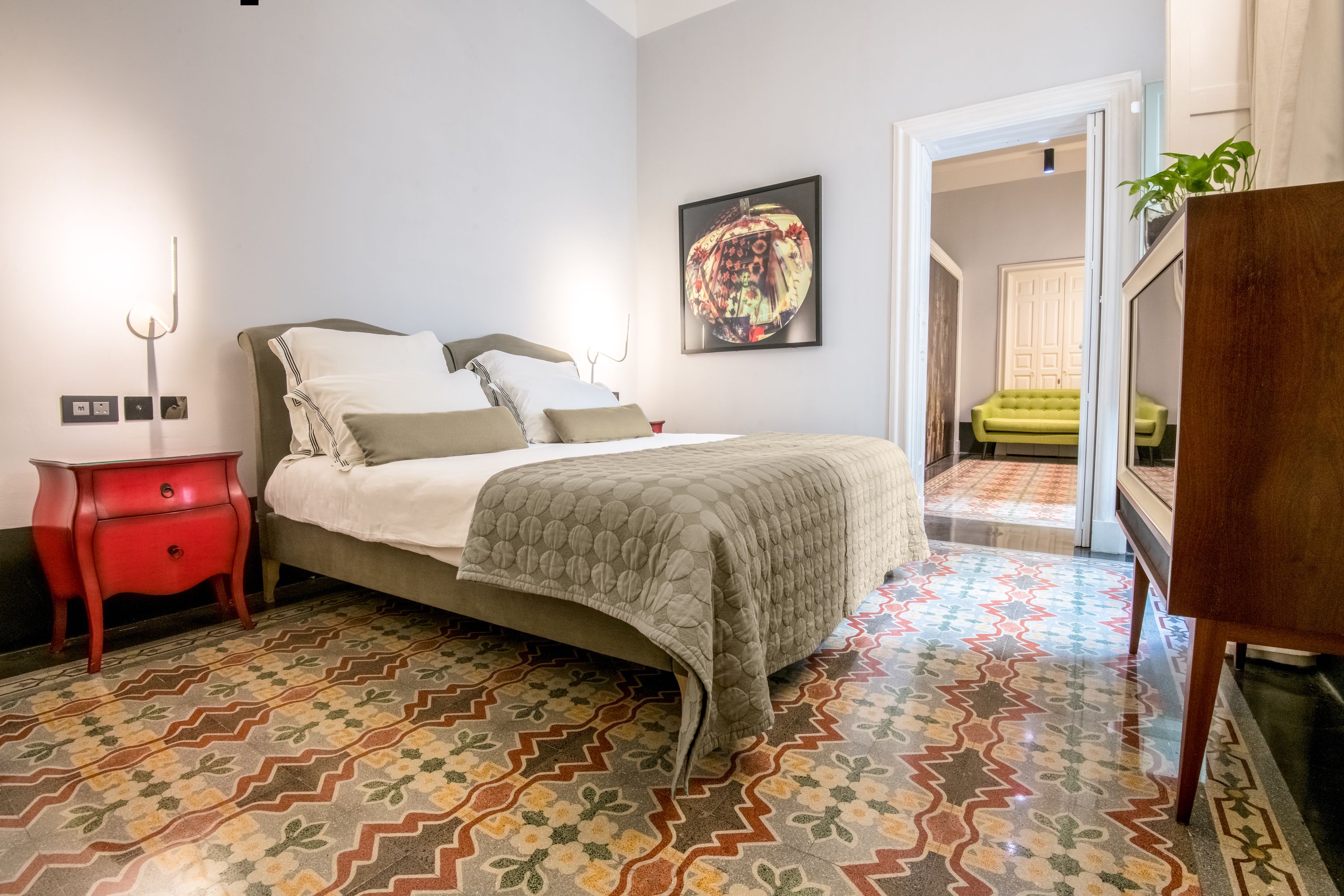 Une chambre à coucher avec un lit, une table de chevet rouge, un sol à motifs et une porte donnant sur une autre pièce.