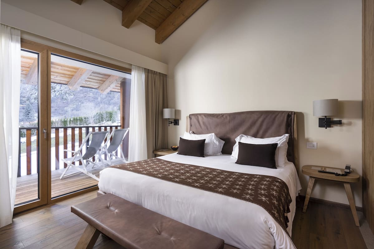 A bedroom with a large bed, a bench, a nightstand, and a view of a balcony with chairs.