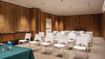 Una sala conferenze con sedie bianche disposte in file, un tavolo color acquamarina con un bicchiere e una parete in legno.