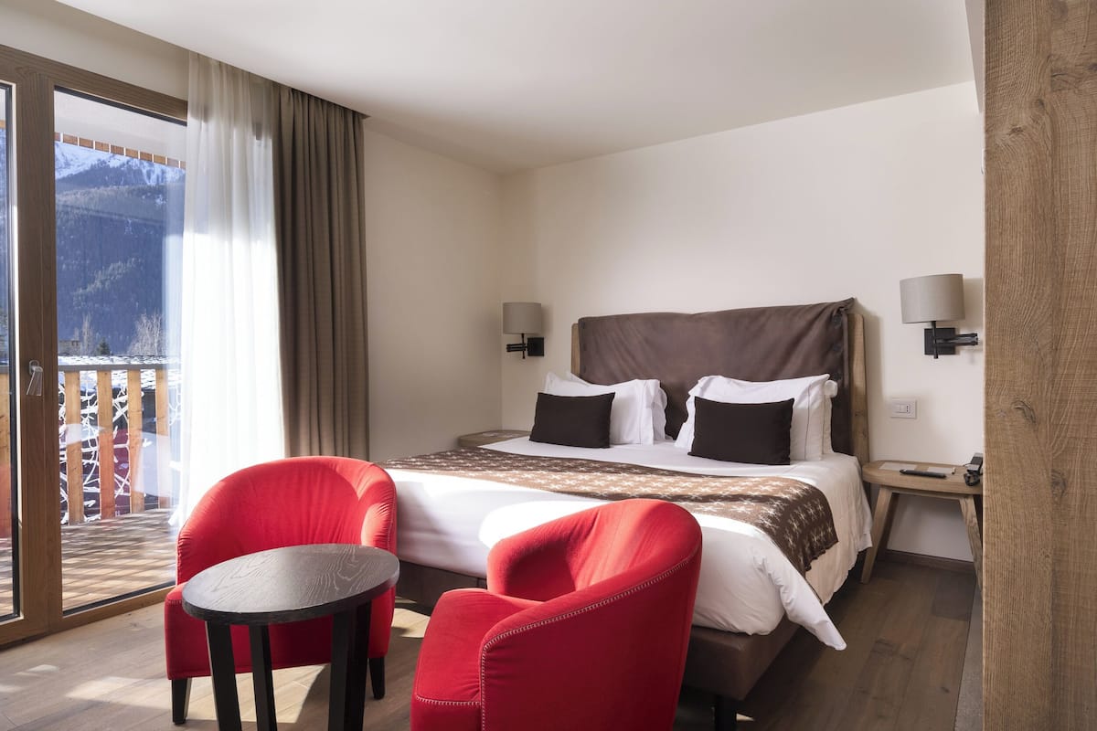 A hotel room with a large bed, two red chairs, a small table, and a balcony with a mountain view.