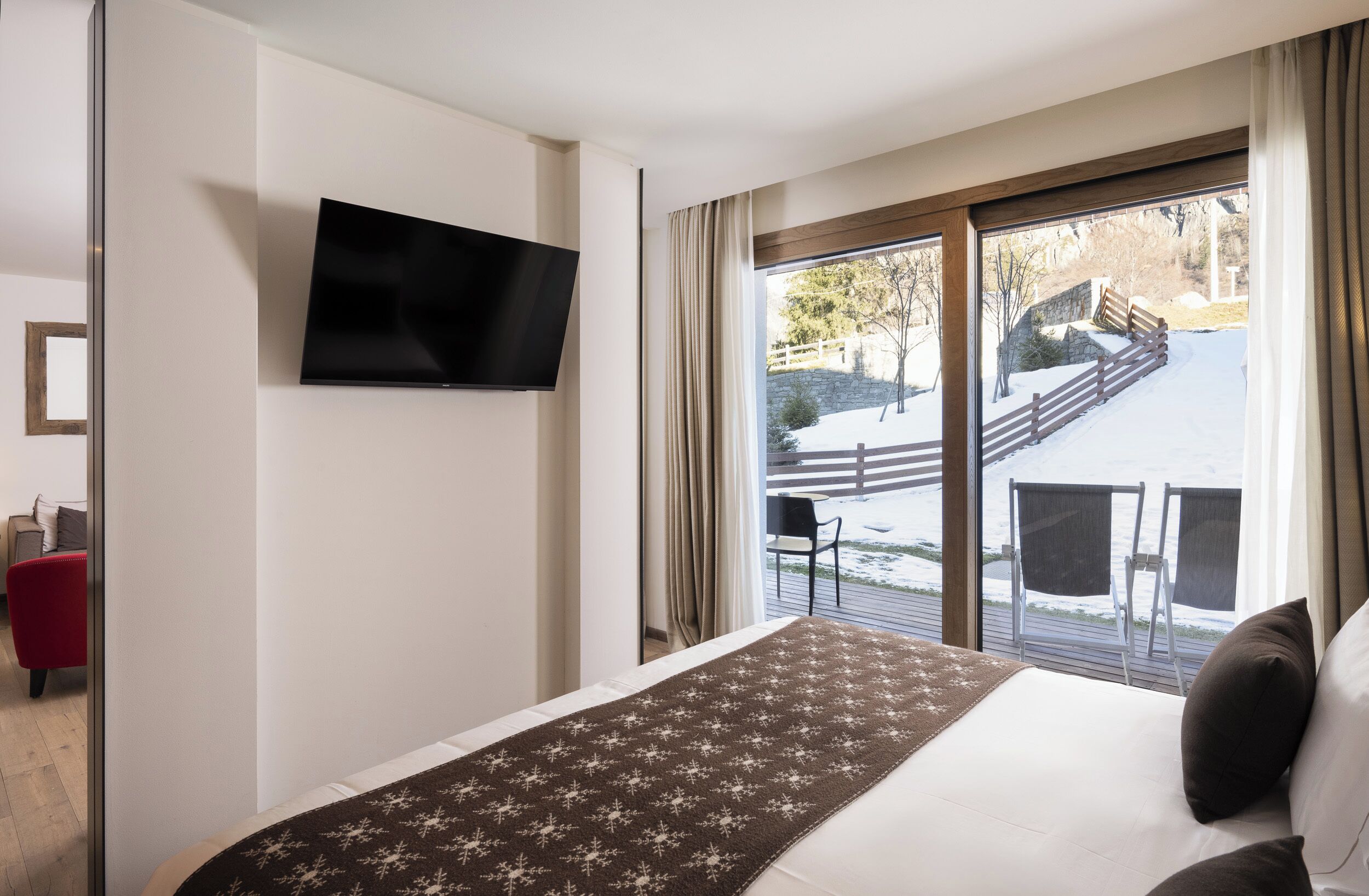 A hotel room with a bed, a TV mounted on the wall, and a view of a snowy outdoor area with a wooden deck and chairs.