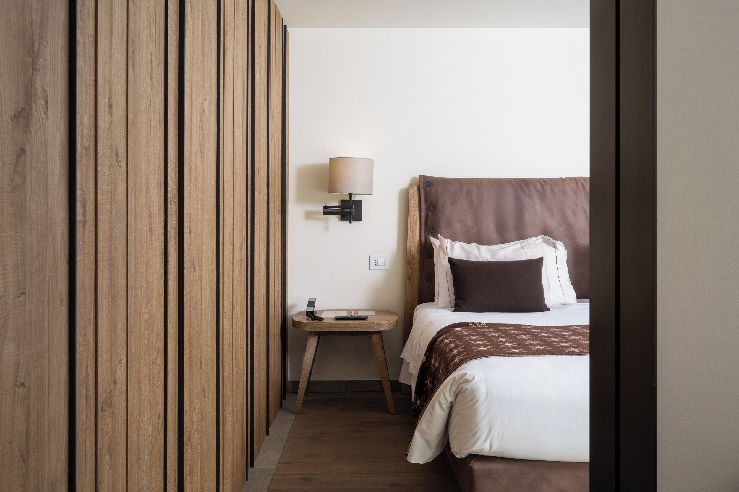 Una habitación de hotel con pared de paneles de madera, una mesita de noche con lámpara, cama con ropa de cama blanca y marrón, y una pequeña mesita.