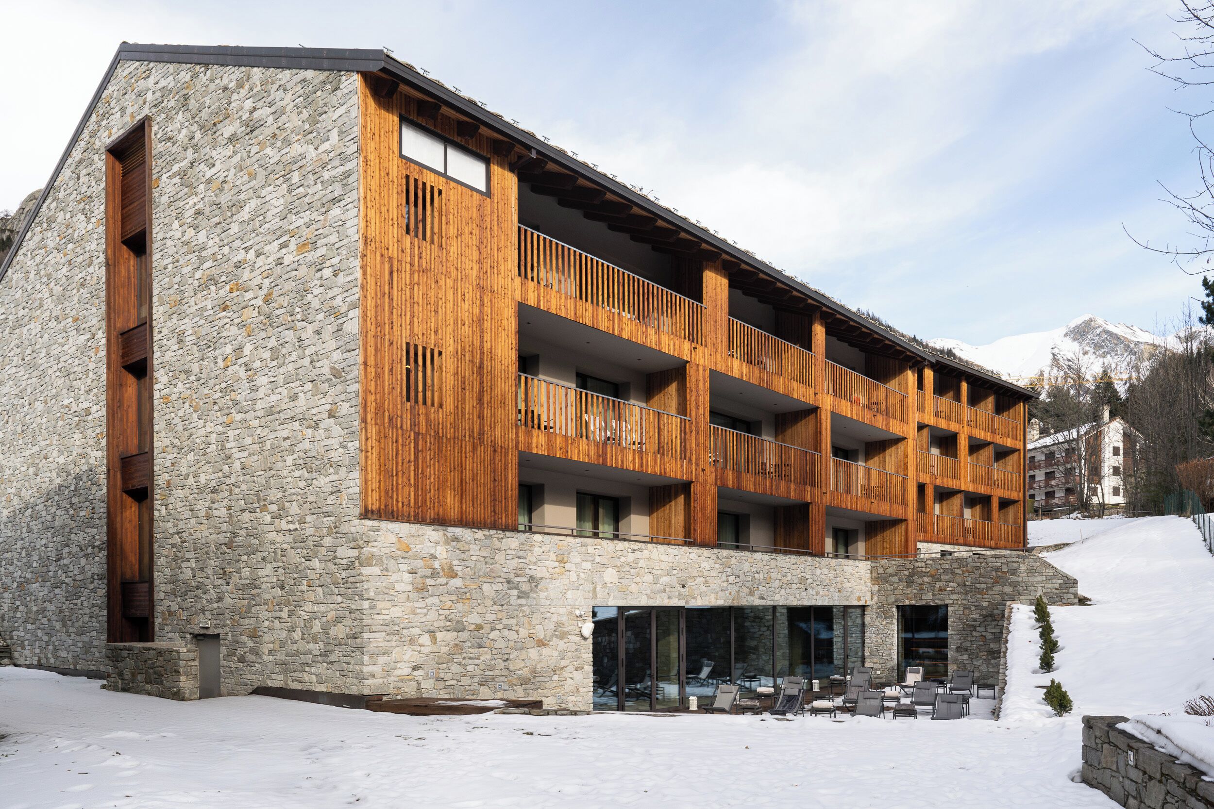 A multi-story building with a stone facade and wooden balconies, set in a snowy landscape with mountains in the background.