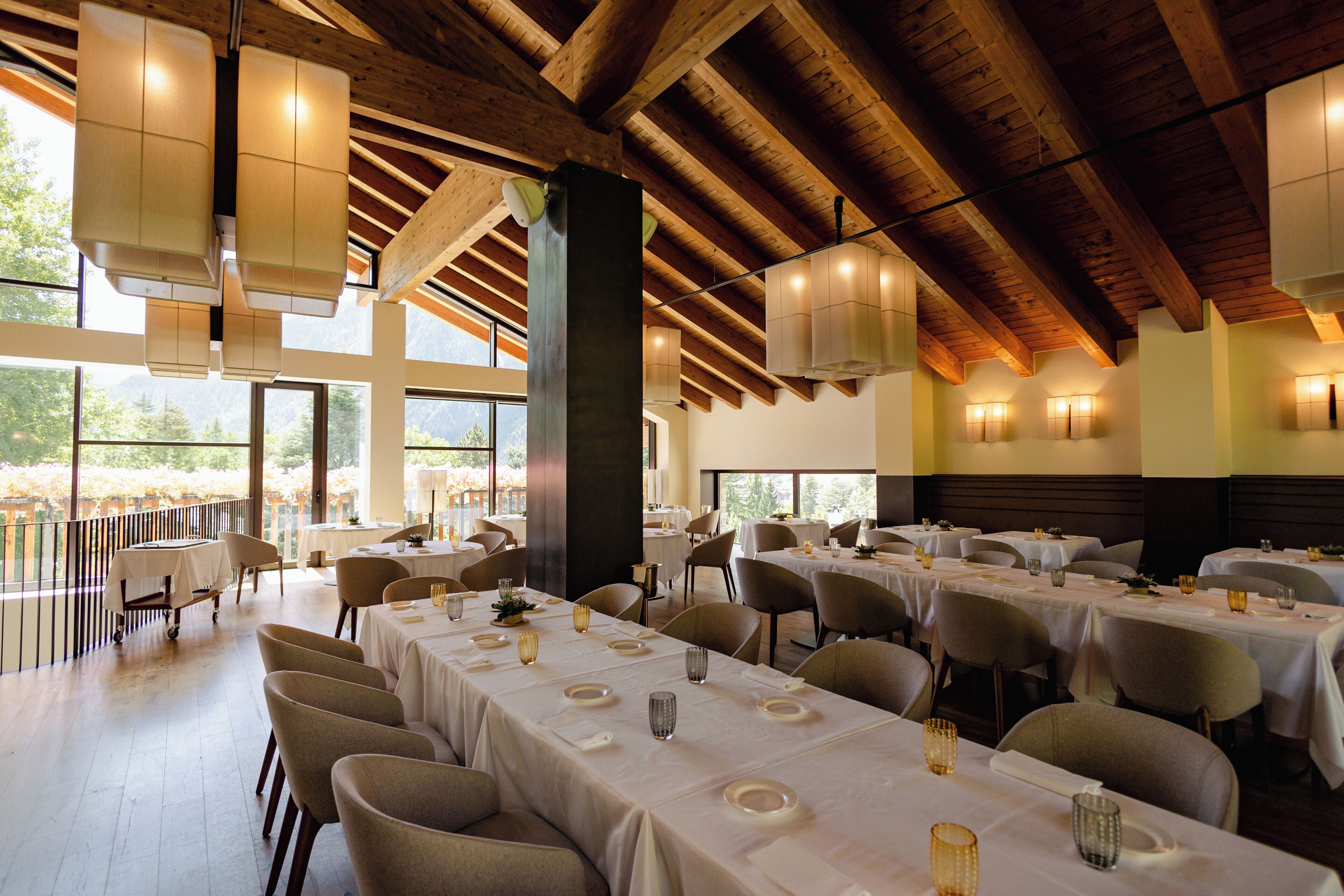 A spacious dining area with multiple tables set for guests, large windows offering views of the outdoors, and a high wooden ceiling.