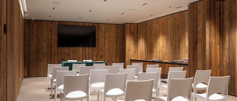 Une salle de conférence avec des chaises blanches disposées en rangées, une table verte et un téléviseur à écran plat fixé au mur.