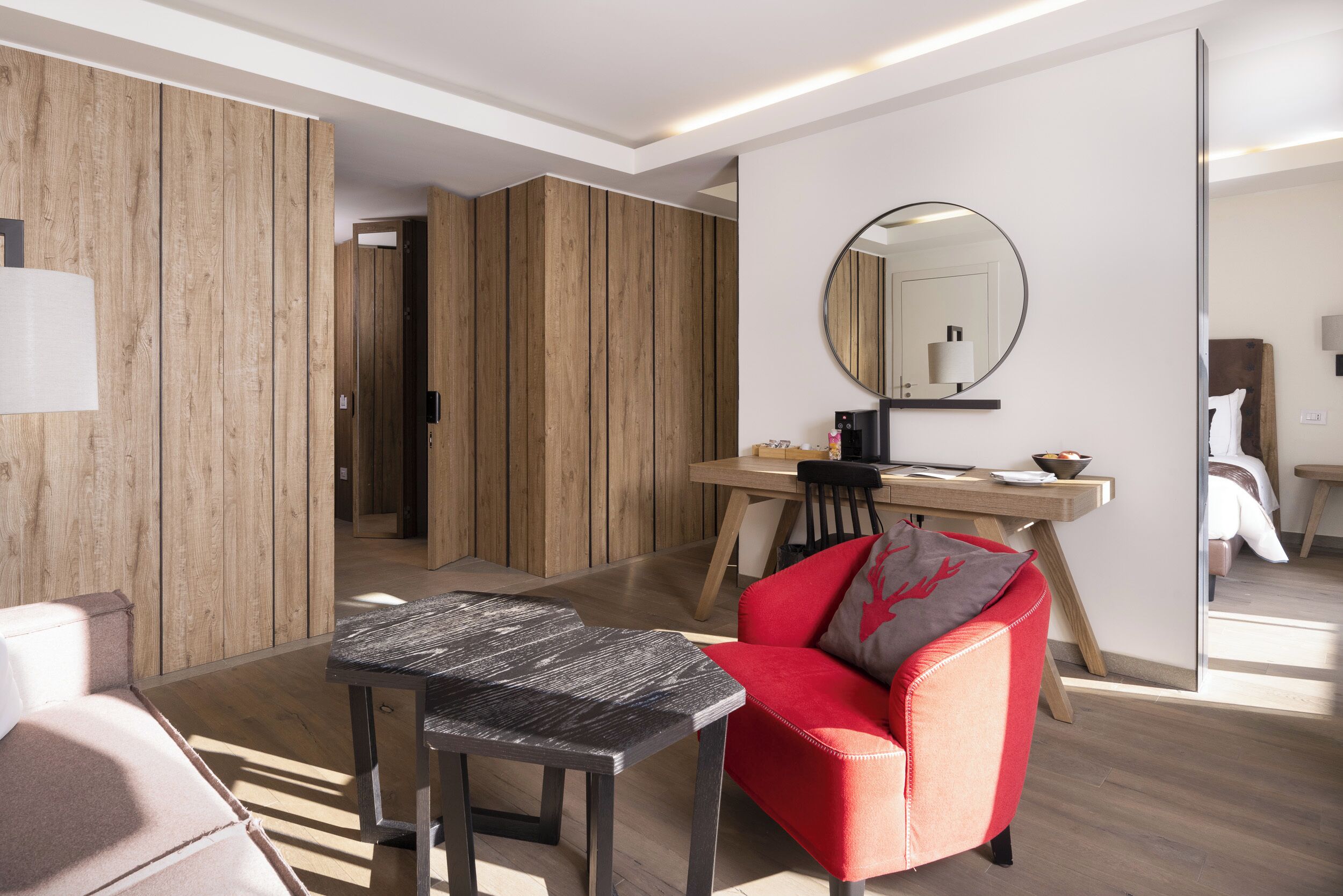 Een moderne hotelkamer met een wand van houten panelen, een ronde spiegel, een eettafel en een rode fauteuil.