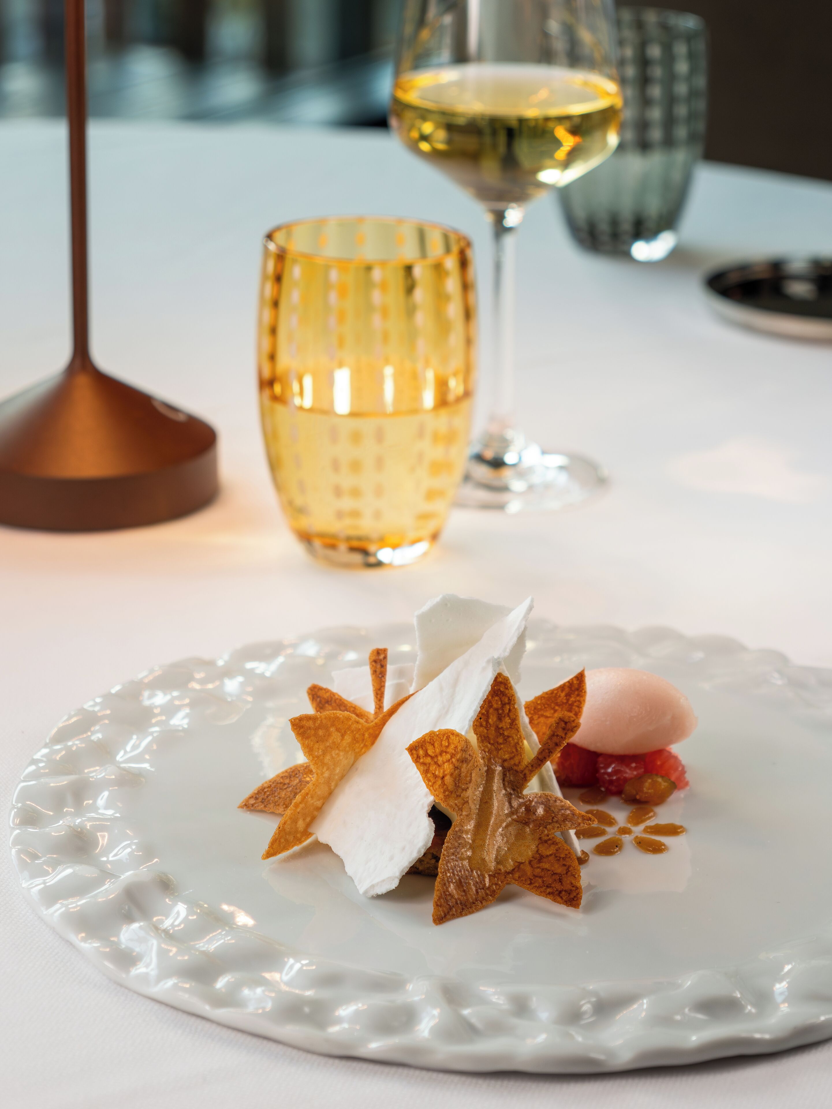 A dessert on a white plate with a textured edge, a glass of white wine, and a candle holder in the background.