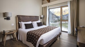 Un dormitorio con una cama grande, cabecera de madera, lámparas de noche, un escritorio y vistas a montañas a través de un ventanal amplio.