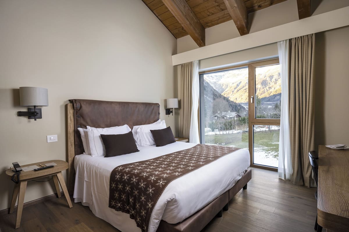 A bedroom with a large bed, wooden headboard, bedside lamps, a desk, and a view of mountains through a large window.