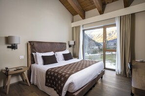 A bedroom with a large bed, wooden headboard, bedside lamps, a desk, and a view of mountains through a large window. - Grand Hotel Courmayeur Montblanc by R Collection Hotels (Courmayeur)