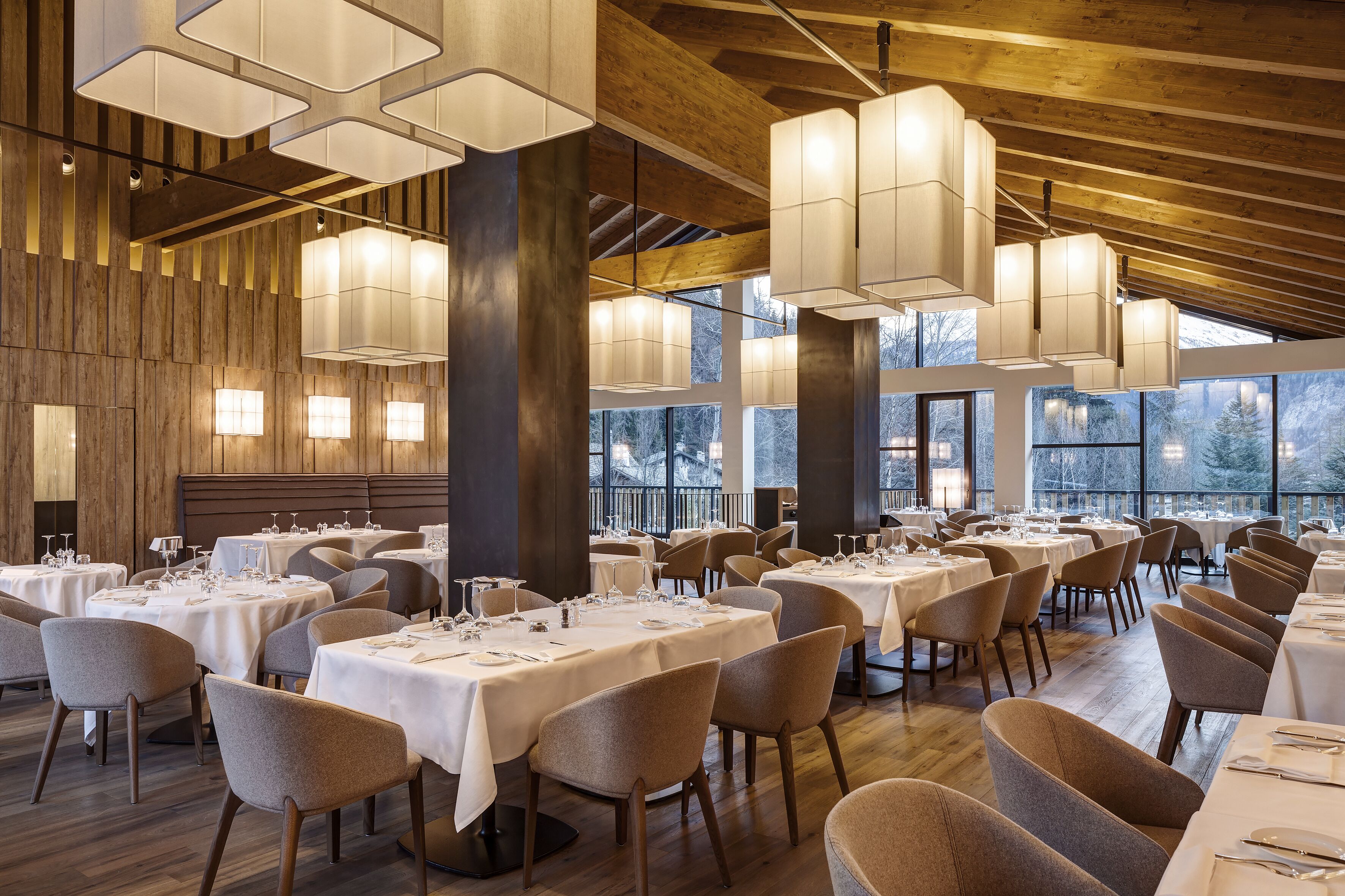 Un restaurante con varias mesas dispuestas para comer, grandes ventanales que ofrecen vistas y moderna iluminación colgante.