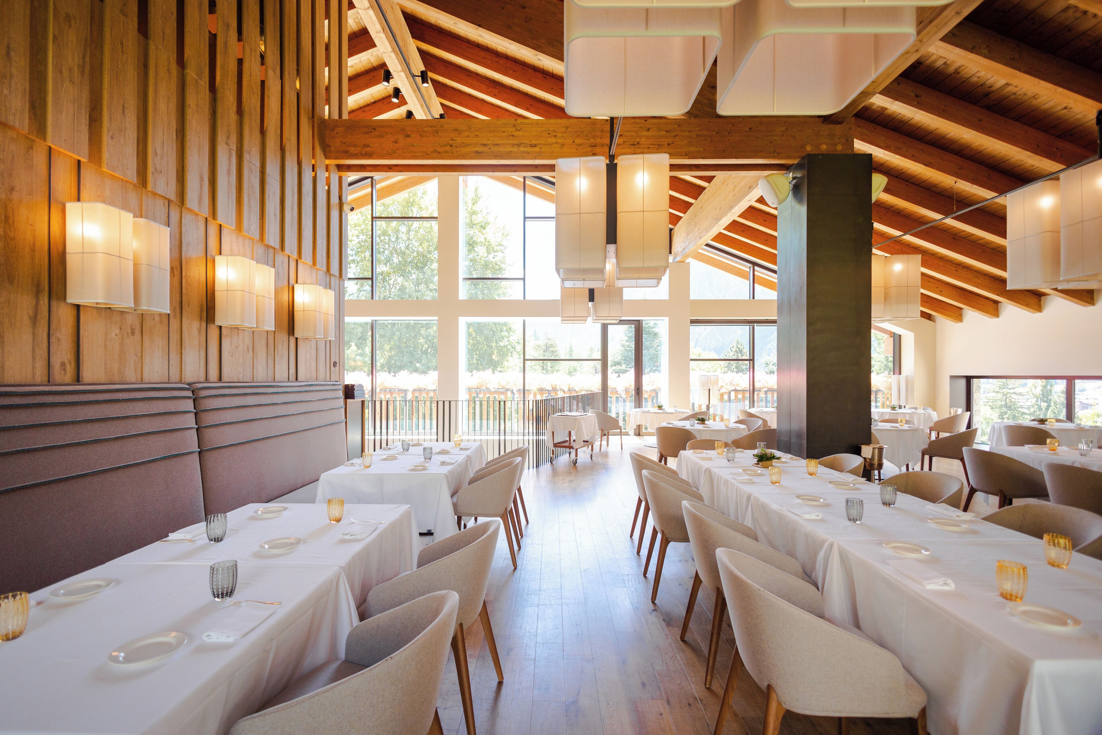 Een restaurant met hoge plafonds, houten balken en grote ramen. Tafels zijn gedekt met witte tafelkleden en stoelen.