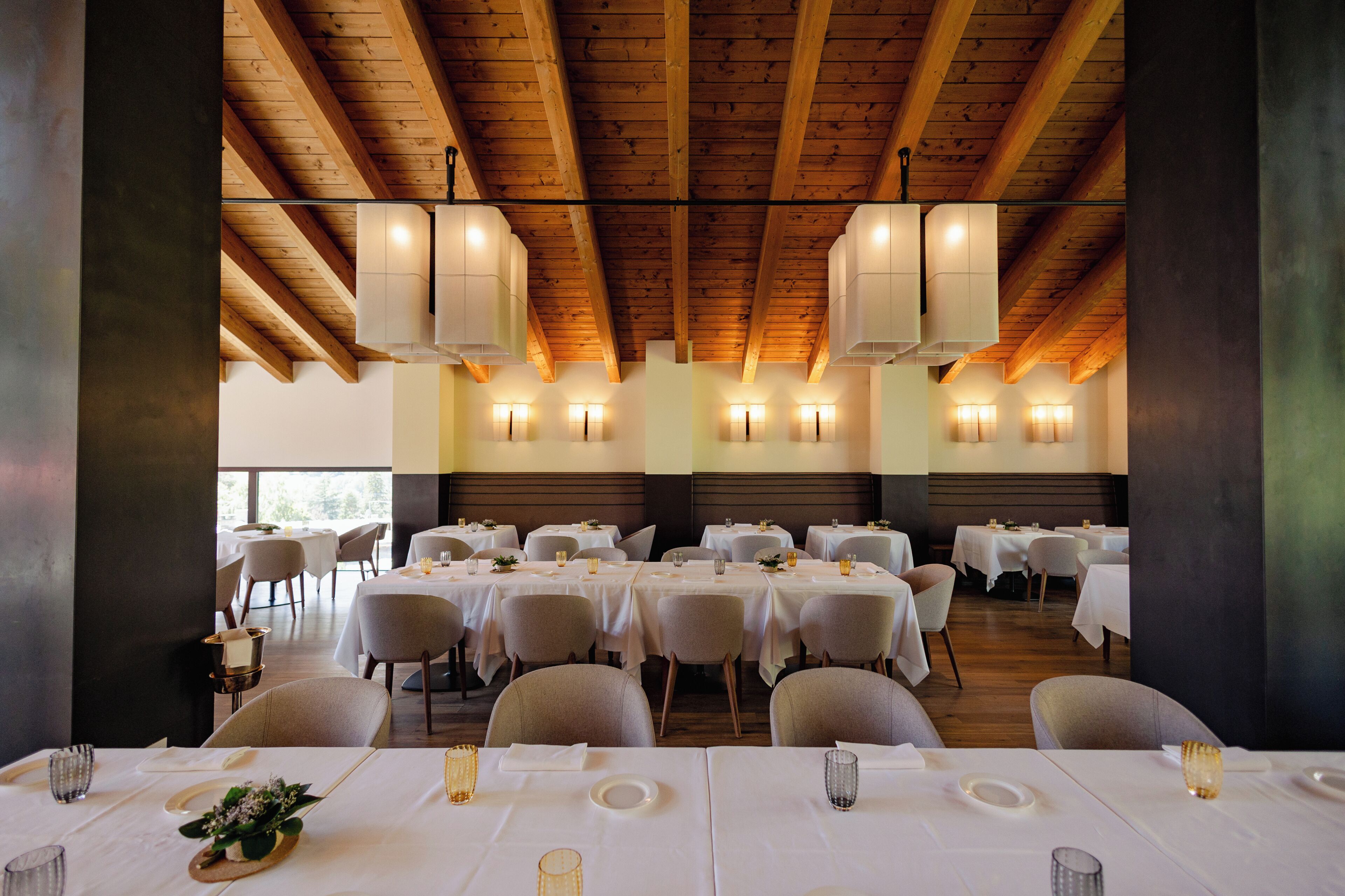 Un restaurant avec de nombreuses tables dressées pour le service, des poutres en bois au plafond et des luminaires fixés aux murs.
