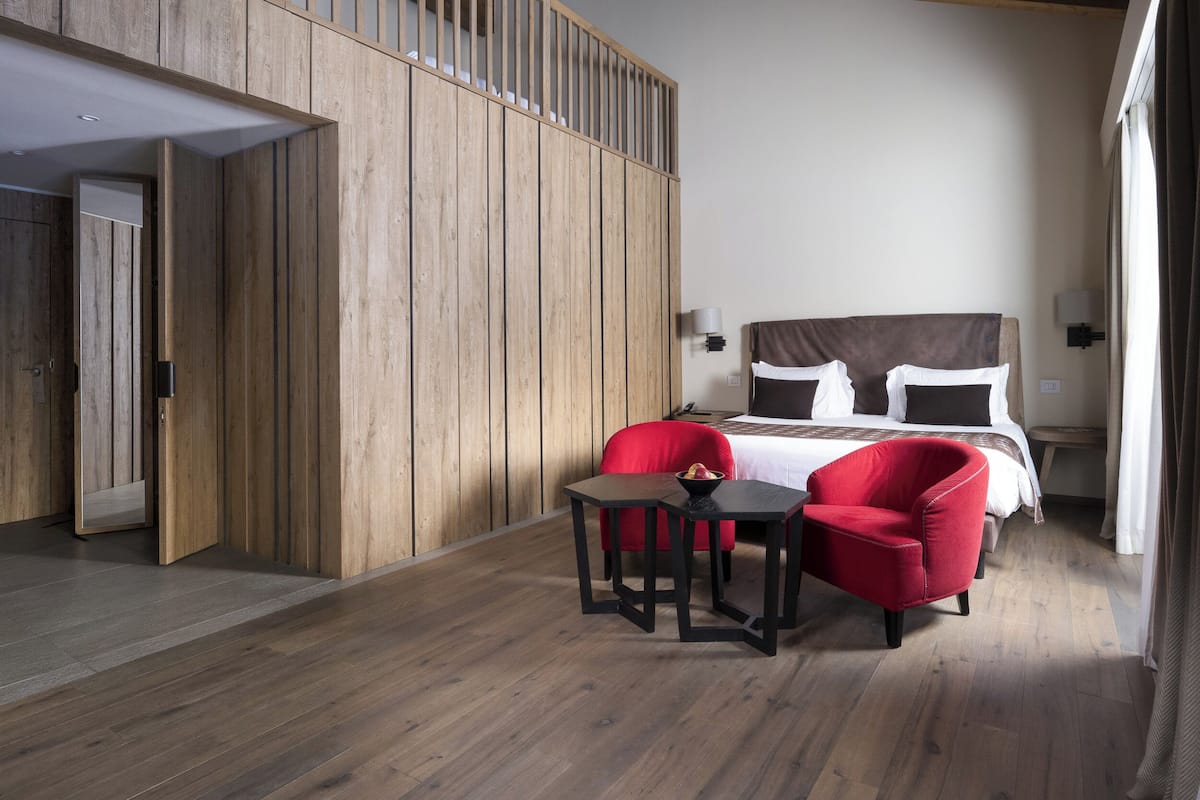 A modern hotel room with a wooden panel wall, a bed with dark bedding, a red chair, and a small table.