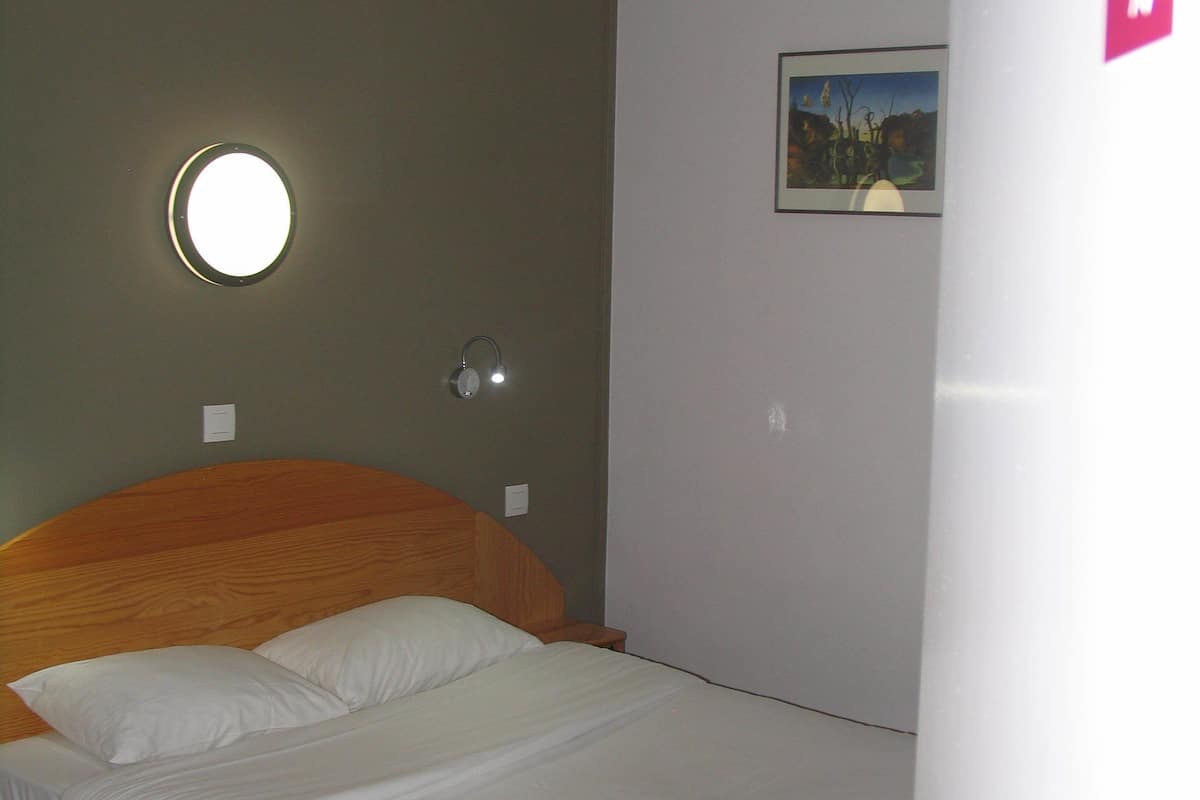 Un lit simple avec une tête de lit en bois, dans une pièce dont le mur est peint dans une couleur claire et qui est agrémentée d’un appliques mural rond.