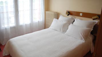 A neatly made bed with white linens in a room with a radiator and a window with sheer curtains.