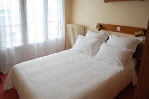 A neatly made bed with white linens in a room with a radiator and a window with sheer curtains. - Hôtel Saint Jean (Bourges)