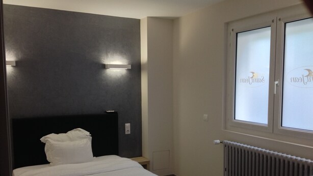 A hotel room with a bed, a nightstand, a window with a view, and a wall-mounted light fixture. - Hôtel Saint Jean (Bourges)