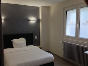 A hotel room with a bed, a nightstand, a window with a view, and a wall-mounted light fixture. - Hôtel Saint Jean (Bourges)