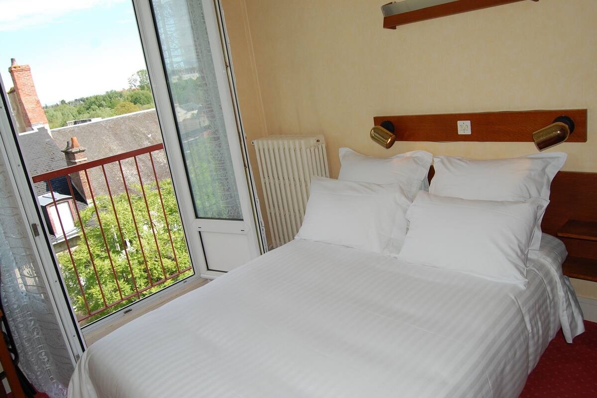 Une chambre d’hôtel avec un grand lit, un radiateur, une fenêtre donnant sur l’extérieur et une tête de lit en bois.