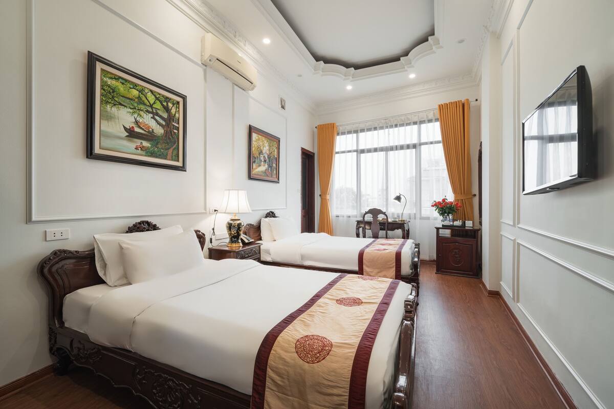 A hotel room with two beds, a TV, a desk with a chair, a window with curtains, and artwork on the walls.
