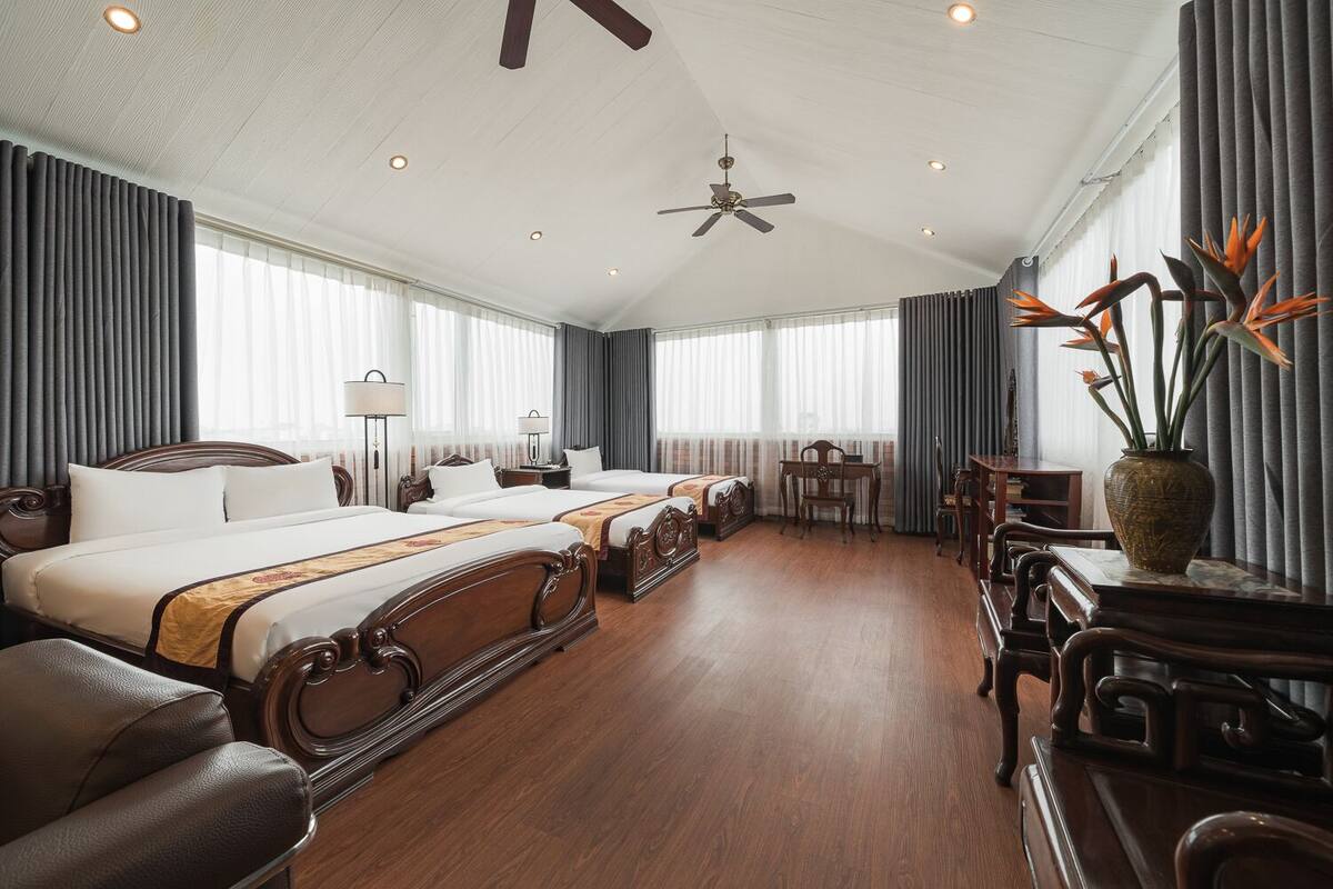 A hotel room with two beds, a desk, a chair, a vase with flowers, and a ceiling fan.