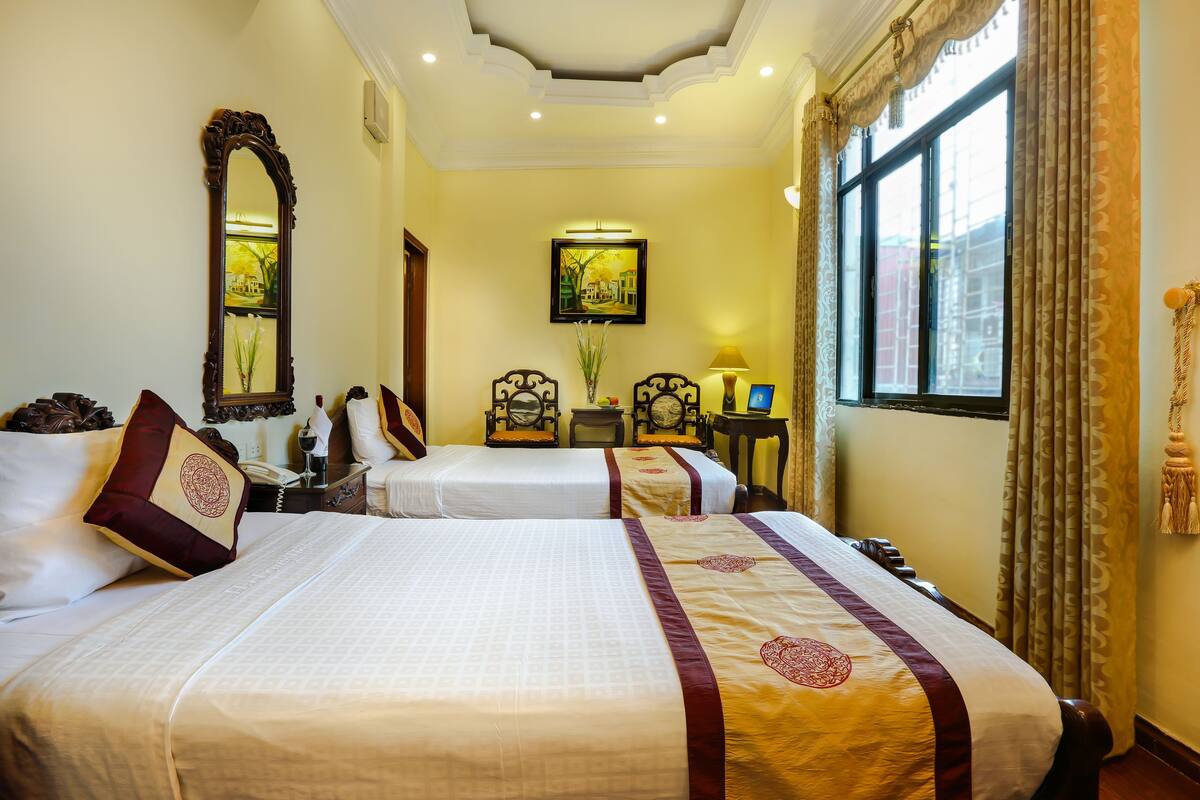 A hotel room with two beds, a large mirror, a painting on the wall, and a window with curtains.
